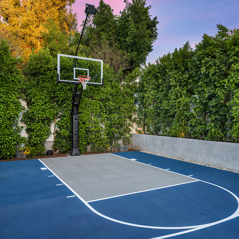 An outdoor basketball court by house with trees, bushes, and fence.